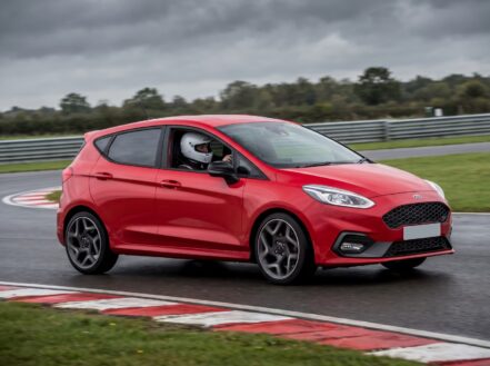 Red Ford Fiesta ST cornering on a British race circuit with red and white kerbing, driver wearing a white full-face racing helmet visible through the windscreen