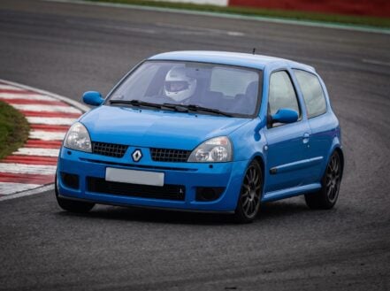 Blue Renault Clio RS at a British race circuit apex with a helmeted driver visible at the wheel, red and white kerbing, overcast trackday light