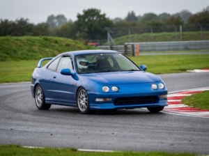 Blue Honda Integra Type R DC2 at a British race circuit apex with helmeted driver visible through windscreen, red and white kerbing, overcast trackday light