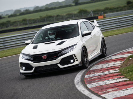 Honda Civic Type R cornering on a British race circuit, driver in a full-face racing helmet visible through the windscreen, red and white kerbing at the apex