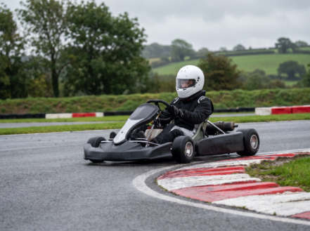 A racing kart cornering at an apex on a British outdoor karting circuit, driver in a full-face helmet visible in the cockpit, red and white kerbing in the foreground