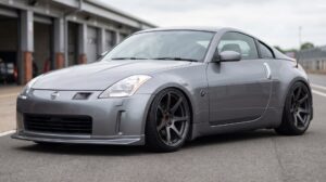 A Nissan 350Z slammed on coilovers in a pit lane. Dark aftermarket wheels, open hub with wheel nuts showing, no centre cap.
