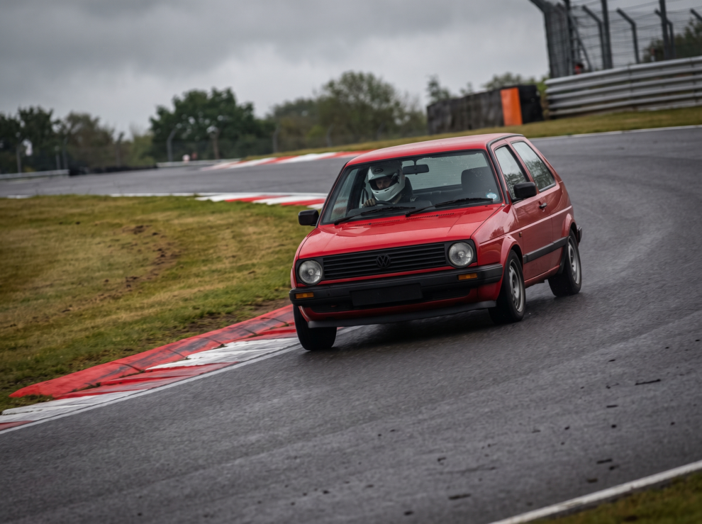 Red Volkswagen Golf Mk2 at a British race circuit apex with helmeted driver visible, red and white kerbing, overcast trackday light